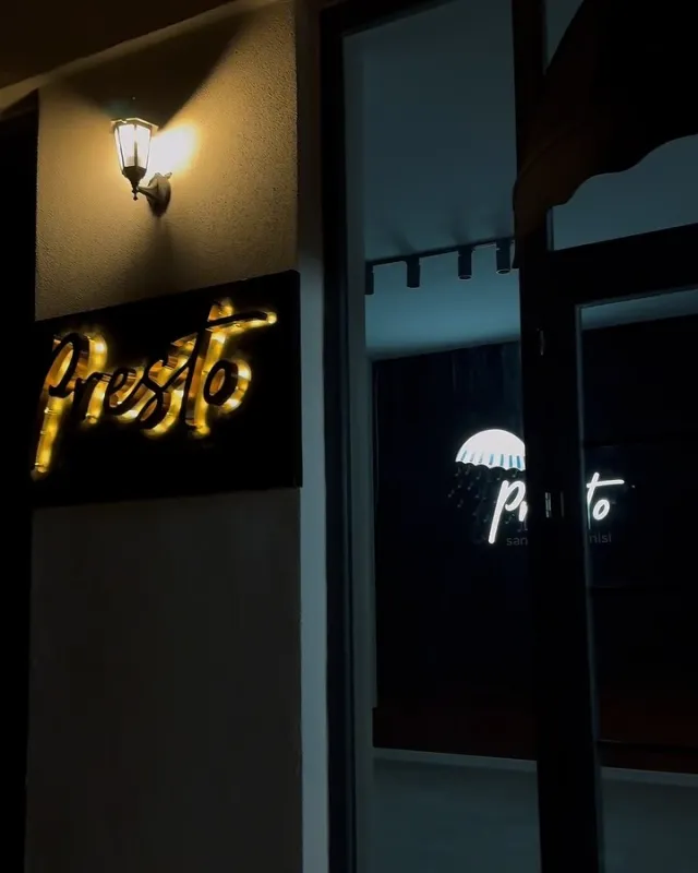 Presto girişinde siyah pano üzerinde altın ışıklı yazı ve cam kapıda beyaz logo